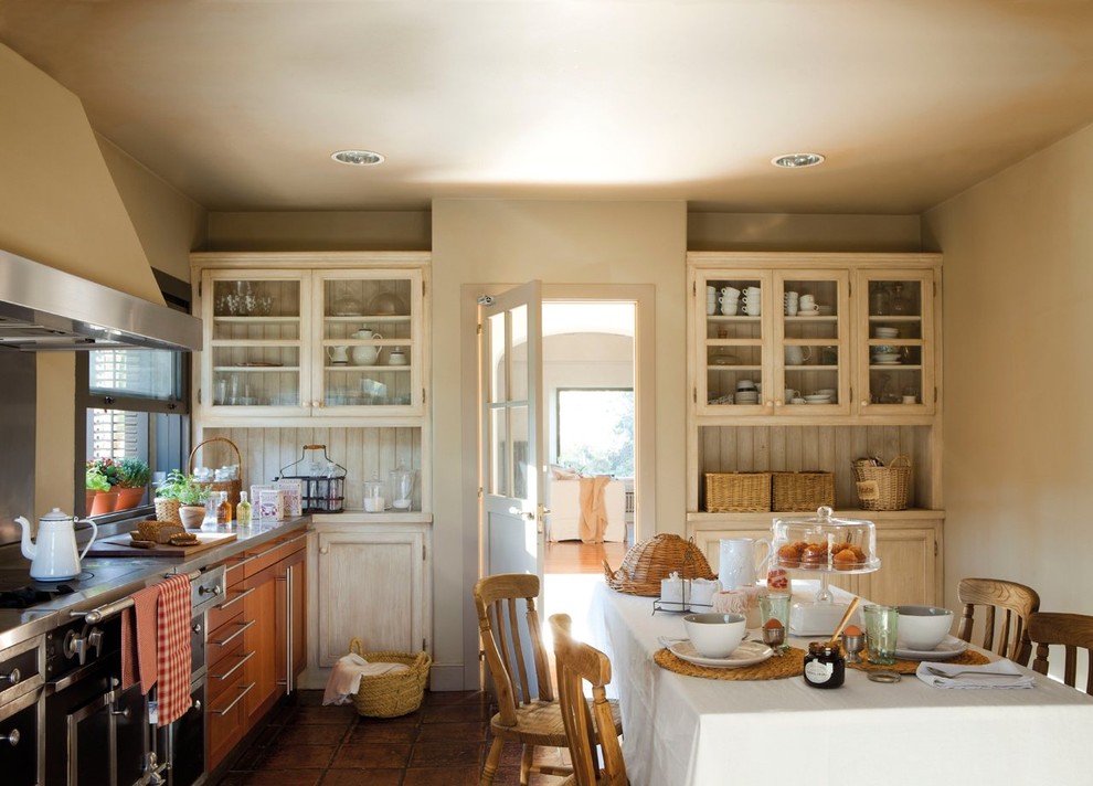 Design ideas for a mid-sized country l-shaped eat-in kitchen in Other with glass-front cabinets, black appliances, terra-cotta floors, white cabinets and no island.