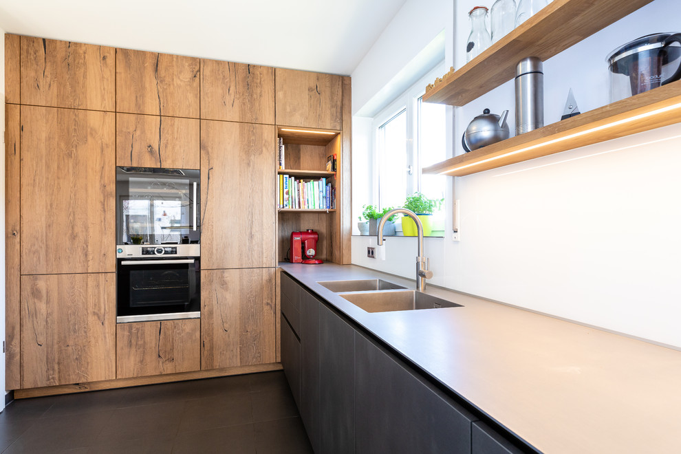 Inspiration for a large contemporary l-shaped open plan kitchen in Frankfurt with an integrated sink, flat-panel cabinets, stainless steel benchtops, black appliances, with island, black floor, grey benchtop and light wood cabinets.