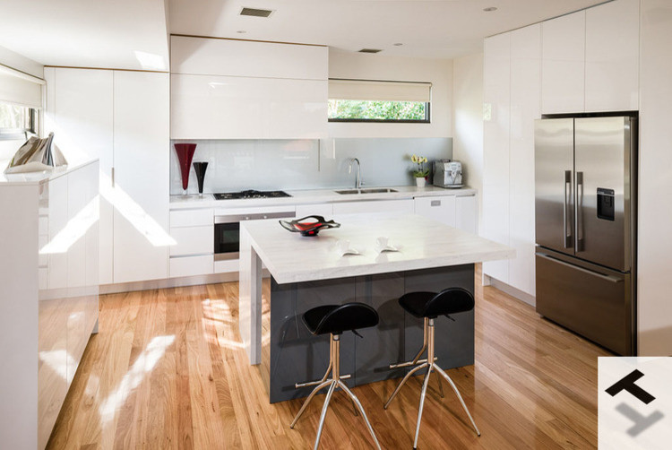 This is an example of a modern kitchen in Melbourne with an integrated sink, white cabinets, solid surface benchtops, metallic splashback, glass sheet splashback, black appliances and light hardwood floors.