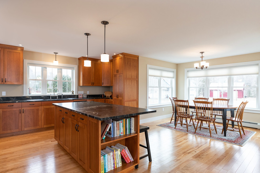 Design ideas for a large country l-shaped eat-in kitchen in Portland Maine with an undermount sink, shaker cabinets, medium wood cabinets, granite benchtops, stainless steel appliances, light hardwood floors, with island, brown floor and black benchtop.