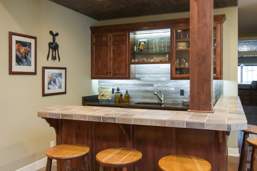 Photo of a mid-sized country u-shaped seated home bar in Kansas City with an undermount sink, raised-panel cabinets, dark wood cabinets, tile benchtops, grey splashback, timber splashback, medium hardwood floors and brown floor.
