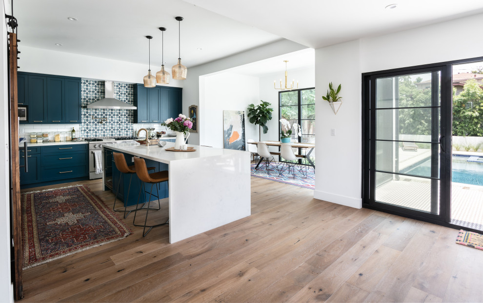 Photo of a mid-sized mediterranean l-shaped eat-in kitchen in Los Angeles with a farmhouse sink, shaker cabinets, blue cabinets, quartz benchtops, white splashback, ceramic splashback, stainless steel appliances, medium hardwood floors, with island, brown floor and white benchtop.