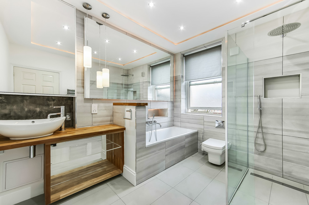 Photo of a large modern kids wet room bathroom in London with glass-front cabinets, grey cabinets, a drop-in tub, a wall-mount toilet, gray tile, ceramic tile, grey walls, porcelain floors, a vessel sink, wood benchtops, grey floor, an open shower and brown benchtops.