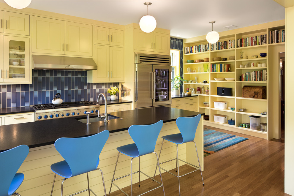 This is an example of a large scandinavian l-shaped eat-in kitchen in Portland with a farmhouse sink, glass-front cabinets, yellow cabinets, granite benchtops, blue splashback, ceramic splashback, stainless steel appliances, medium hardwood floors, with island and black benchtop.