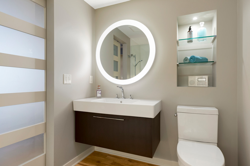 This is an example of a small modern 3/4 bathroom in Minneapolis with flat-panel cabinets, brown cabinets, a curbless shower, a one-piece toilet, grey walls, vinyl floors, a wall-mount sink, brown floor, an open shower and white benchtops.