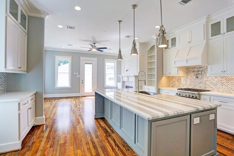 Mid-sized traditional galley open plan kitchen in Houston with a single-bowl sink, recessed-panel cabinets, white cabinets, granite benchtops, grey splashback, glass tile splashback, stainless steel appliances, plywood floors, with island and brown floor.