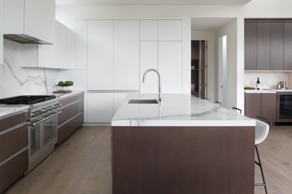 This is an example of a modern kitchen in San Francisco with flat-panel cabinets, white cabinets, white splashback, panelled appliances, light hardwood floors, with island, brown floor and white benchtop.