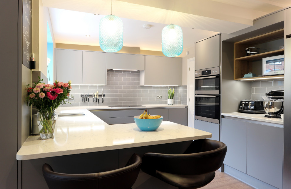 This is an example of a large contemporary grey and teal u-shaped kitchen/diner in Hertfordshire with an integrated sink, flat-panel cabinets, grey cabinets, quartz worktops, grey splashback, metro tiled splashback, stainless steel appliances, lino flooring, a breakfast bar, brown floors, white worktops and feature lighting.