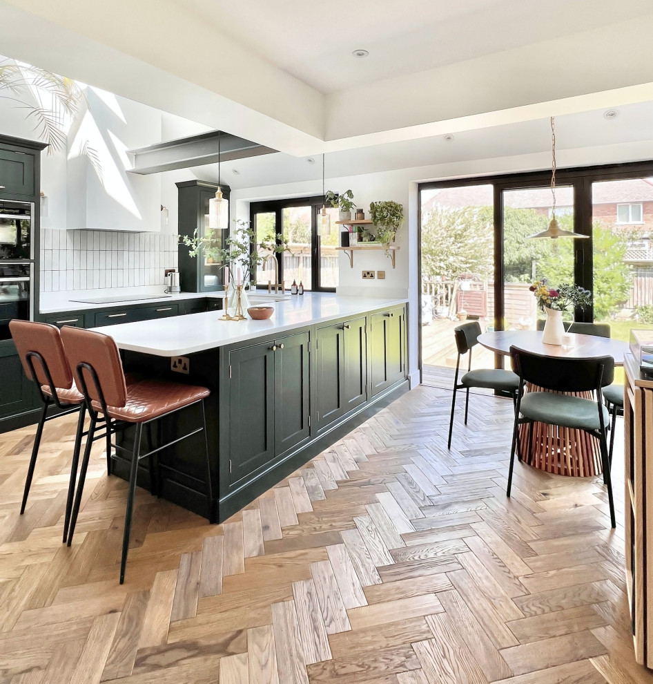 Bohemian u-shaped kitchen with a belfast sink, shaker cabinets, green cabinets, quartz worktops, white splashback, porcelain splashback, stainless steel appliances, medium hardwood flooring, an island, brown floors, white worktops and exposed beams.