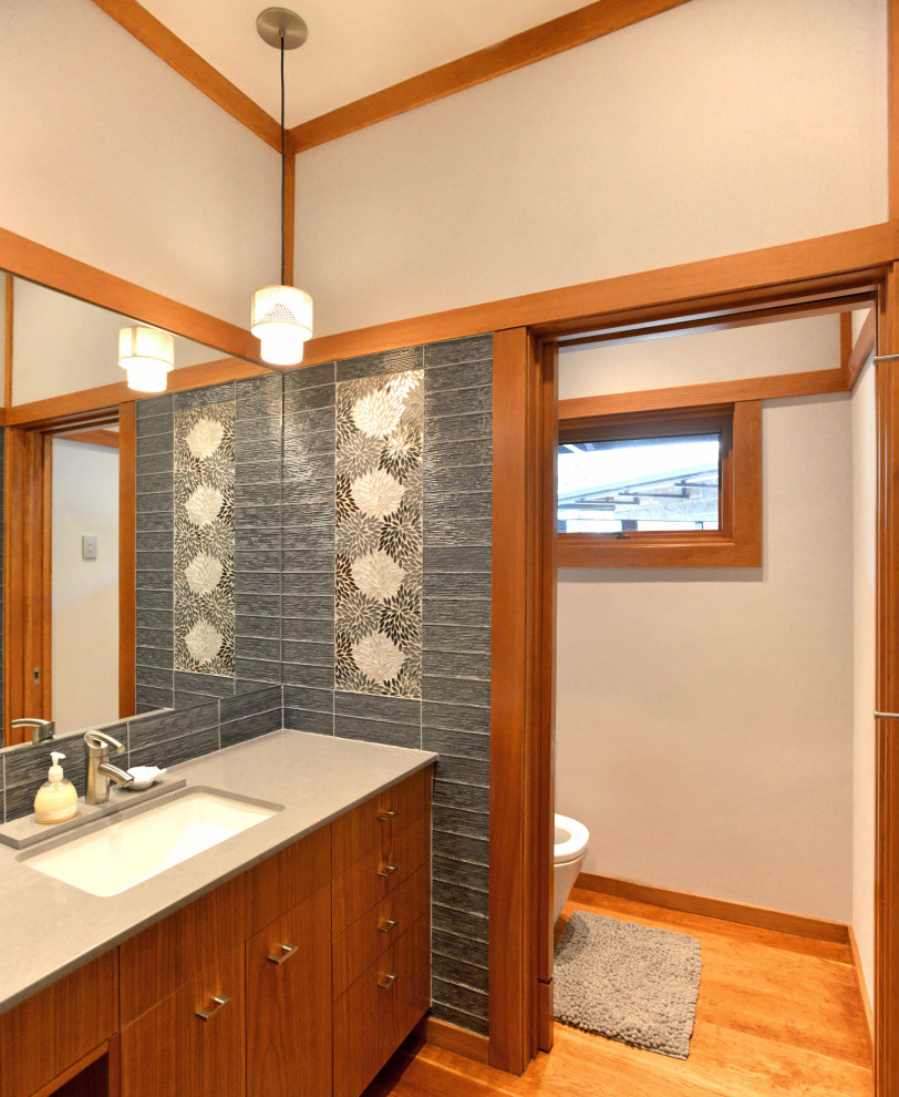 Photo of a mid-sized asian bathroom in Austin with flat-panel cabinets, grey cabinets, a one-piece toilet, beige tile, glass tile, engineered quartz benchtops, beige benchtops, a single vanity, a floating vanity, beige walls, medium hardwood floors, a drop-in sink, brown floor and an enclosed toilet.