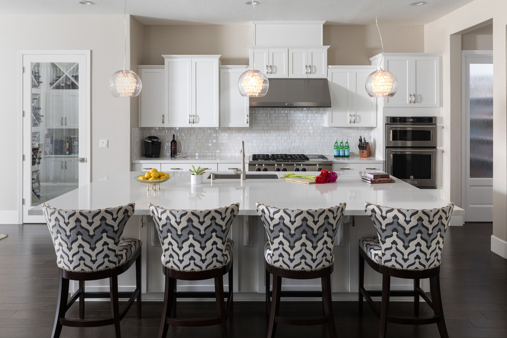 This is an example of a large transitional single-wall eat-in kitchen in San Francisco with an undermount sink, shaker cabinets, solid surface benchtops, white splashback, glass tile splashback, stainless steel appliances, dark hardwood floors, with island, brown floor and white benchtop.