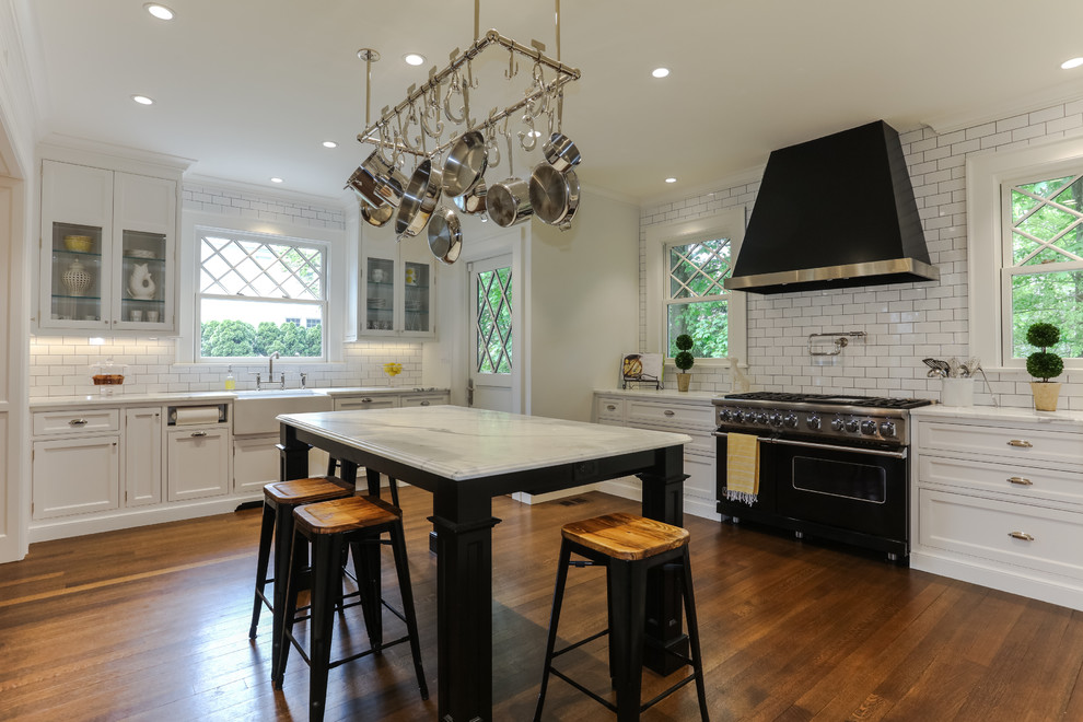 Example of a transitional l-shaped medium tone wood floor and brown floor kitchen design with a farmhouse sink, shaker cabinets, white cabinets, white backsplash, subway tile backsplash, black appliances, an island and white countertops