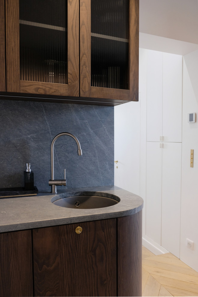 Photo of a small contemporary galley open plan kitchen in Other with an undermount sink, glass-front cabinets, dark wood cabinets, granite benchtops, grey splashback, granite splashback, panelled appliances, light hardwood floors, grey benchtop and exposed beam.