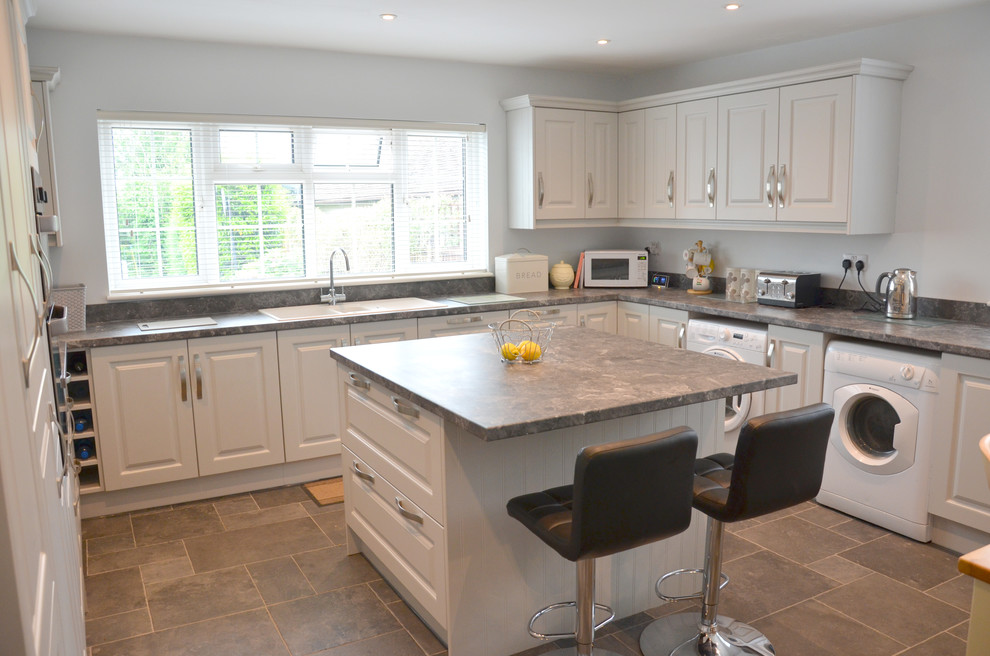 Mid-sized traditional u-shaped eat-in kitchen in Essex with a double-bowl sink, raised-panel cabinets, grey cabinets, laminate benchtops, grey splashback, glass sheet splashback, porcelain floors, with island, grey floor and grey benchtop.
