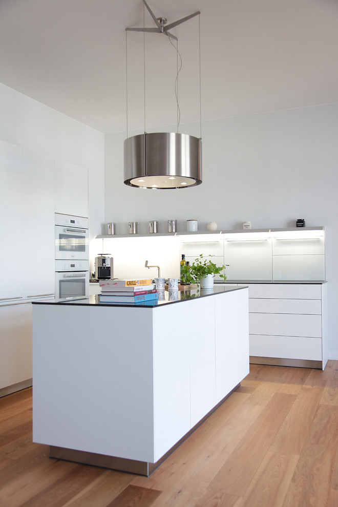 Design ideas for a mid-sized contemporary l-shaped open plan kitchen in Hamburg with flat-panel cabinets, white cabinets, glass benchtops, white splashback, glass sheet splashback, white appliances, medium hardwood floors, with island, brown floor and black benchtop.