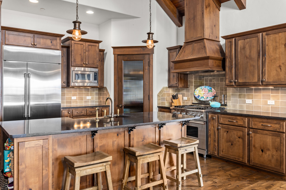Mid-sized mountain style l-shaped dark wood floor, brown floor and wood ceiling open concept kitchen photo with a farmhouse sink, shaker cabinets, brown cabinets, quartz countertops, gray backsplash, ceramic backsplash, stainless steel appliances, an island and brown countertops