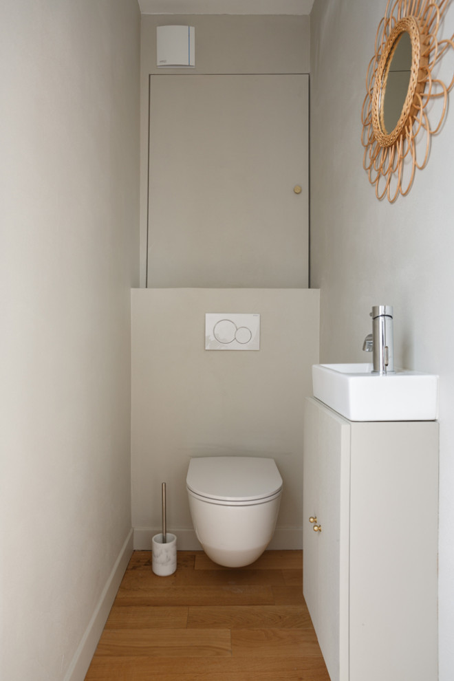 Mid-sized powder room in London with flat-panel cabinets, beige cabinets, a wall-mount toilet, beige tile, beige walls, light hardwood floors, a wall-mount sink, solid surface benchtops, beige floor and white benchtops.
