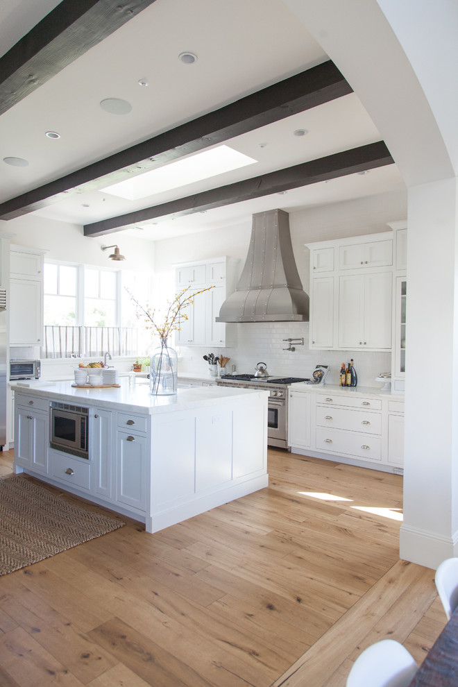 Large transitional l-shaped light wood floor eat-in kitchen photo in Phoenix with a farmhouse sink, white cabinets, white backsplash, stainless steel appliances, an island, stainless steel countertops, shaker cabinets and porcelain backsplash