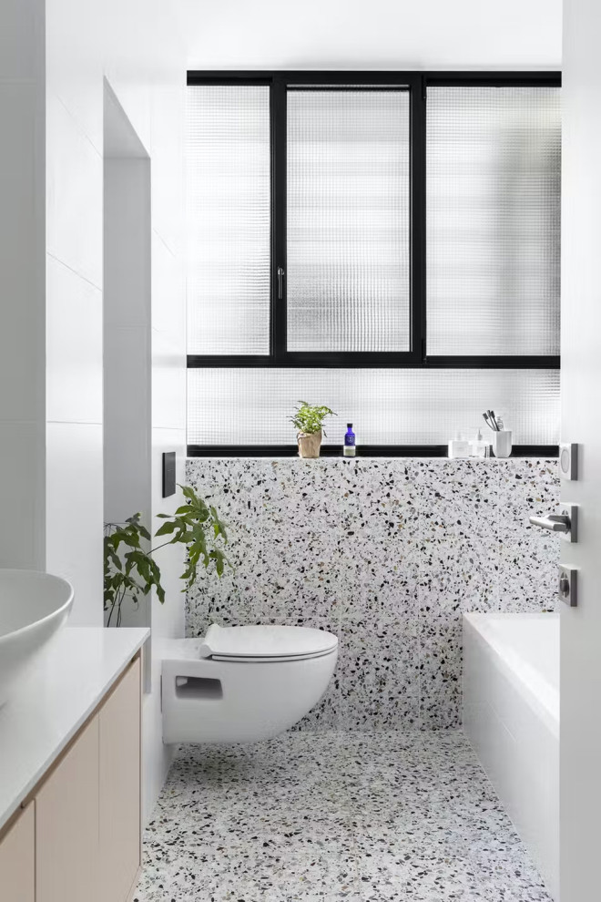 Foto på ett mellanstort vit en-suite badrum, med släta luckor, beige skåp, ett platsbyggt badkar, en dusch/badkar-kombination, en vägghängd toalettstol, vit kakel, marmorkakel, vita väggar, terrazzogolv, ett fristående handfat, marmorbänkskiva, vitt golv och med dusch som är öppen
