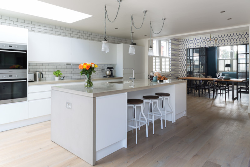 Inspiration for a mid-sized contemporary single-wall open plan kitchen in London with a double-bowl sink, flat-panel cabinets, white cabinets, concrete benchtops, white splashback, ceramic splashback, stainless steel appliances, light hardwood floors and with island.