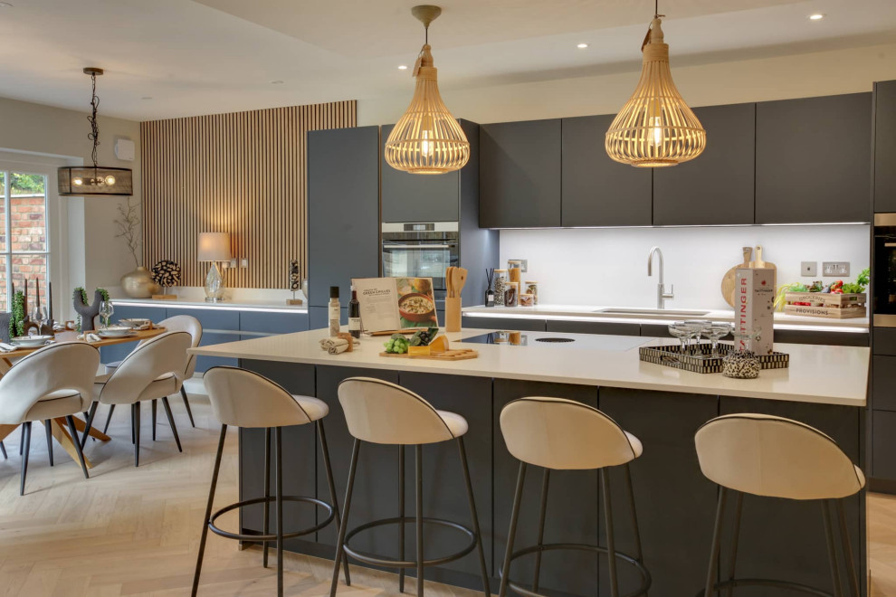 Large contemporary l-shaped open plan kitchen in Hertfordshire with flat-panel cabinets, grey cabinets, quartz worktops, white splashback, black appliances, an island and white worktops.