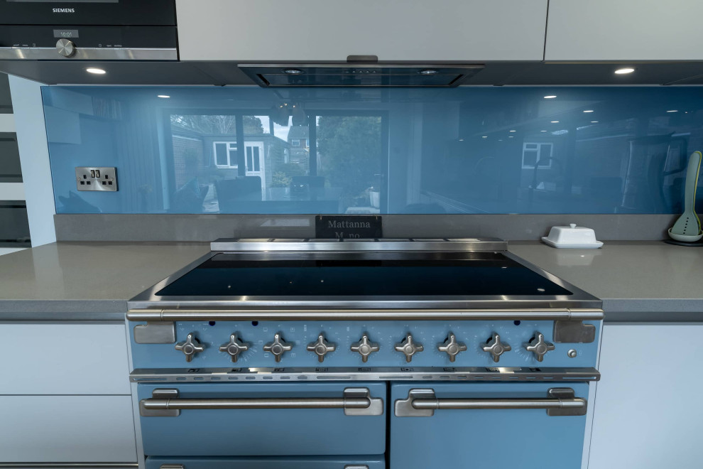 Photo of a medium sized contemporary l-shaped open plan kitchen in Oxfordshire with a single-bowl sink, flat-panel cabinets, blue cabinets, quartz worktops, blue splashback, glass sheet splashback, integrated appliances, an island, multi-coloured floors, grey worktops and feature lighting.