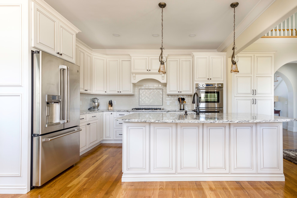 Photo of a mid-sized traditional l-shaped kitchen in Nashville with raised-panel cabinets, white cabinets, quartz benchtops, white splashback, ceramic splashback, stainless steel appliances, medium hardwood floors, with island, brown floor and multi-coloured benchtop.