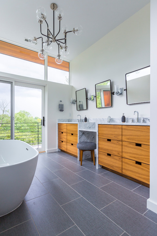 Photo of a contemporary bathroom in Cincinnati with flat-panel cabinets, medium wood cabinets, a freestanding tub, white walls, porcelain floors, an undermount sink, marble benchtops, grey floor and white benchtops.