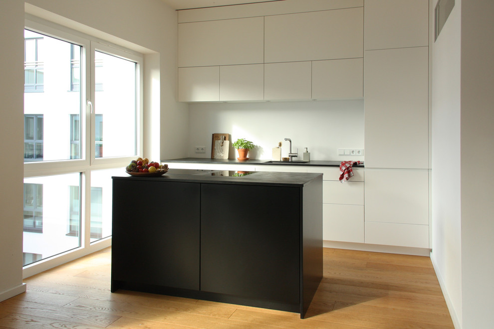 This is an example of a mid-sized modern single-wall open plan kitchen in Berlin with an undermount sink, flat-panel cabinets, white cabinets, granite benchtops, white splashback, slate splashback, black appliances, light hardwood floors, with island and black benchtop.
