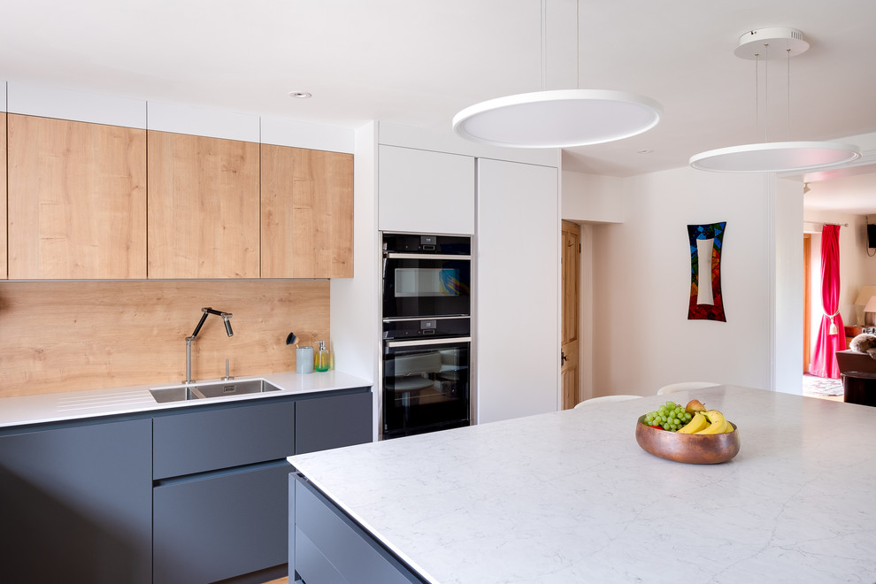 Photo of a mid-sized modern u-shaped open plan kitchen in Gloucestershire with a drop-in sink, flat-panel cabinets, solid surface benchtops, timber splashback, black appliances, with island, beige floor and white benchtop.