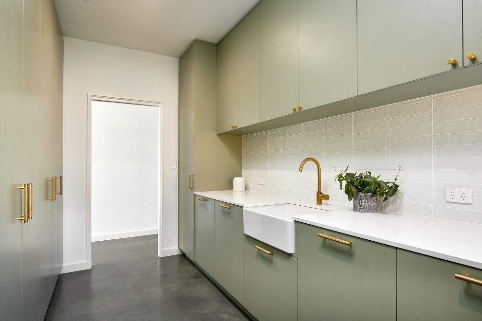 Photo of a large traditional galley dedicated laundry room in Perth with a farmhouse sink, flat-panel cabinets, green cabinets, quartz benchtops, white splashback, subway tile splashback, white walls, concrete floors, a stacked washer and dryer, grey floor, white benchtop and brick walls.