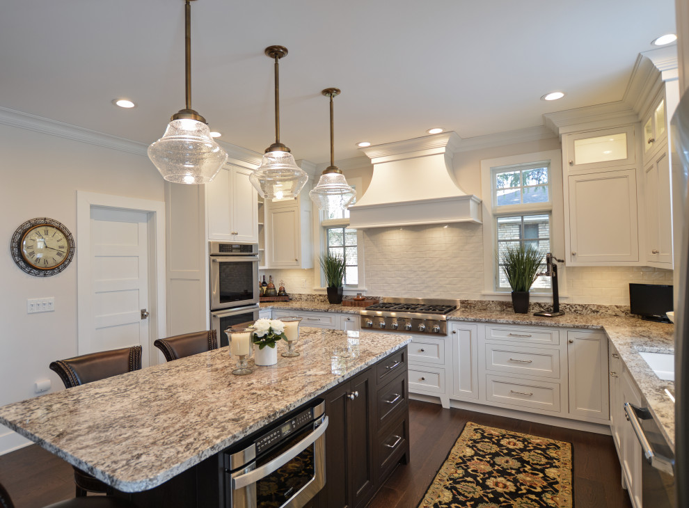 Example of a transitional dark wood floor and brown floor eat-in kitchen design with an undermount sink, shaker cabinets, white cabinets, granite countertops, white backsplash, ceramic backsplash, stainless steel appliances, an island and beige countertops