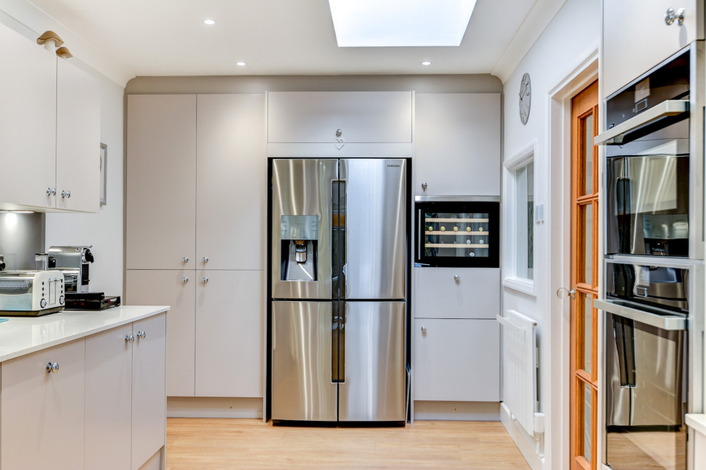 Photo of a mid-sized contemporary galley eat-in kitchen in Sussex with an integrated sink, flat-panel cabinets, grey cabinets, quartzite benchtops, grey splashback, glass tile splashback, black appliances, medium hardwood floors, no island, brown floor and white benchtop.