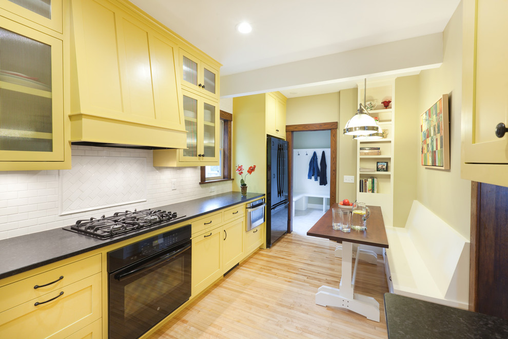 Photo of a mid-sized traditional u-shaped eat-in kitchen in Minneapolis with a farmhouse sink, flat-panel cabinets, yellow cabinets, granite benchtops, white splashback, ceramic splashback, black appliances, light hardwood floors and no island.