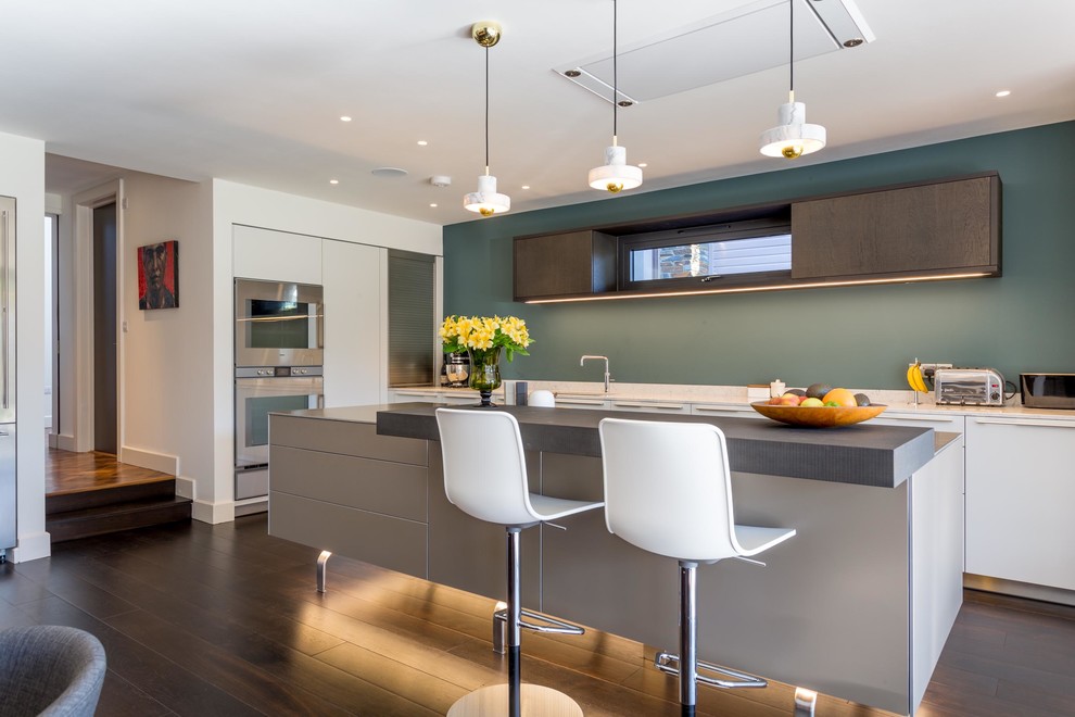 Inspiration for a mid-sized contemporary galley eat-in kitchen in Devon with flat-panel cabinets, stainless steel benchtops, panelled appliances, with island, beige splashback, marble splashback, dark hardwood floors and brown floor.