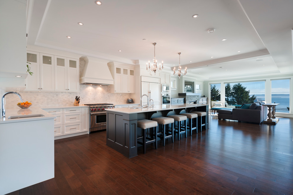 Design ideas for an expansive transitional l-shaped open plan kitchen in Vancouver with an undermount sink, shaker cabinets, white cabinets, marble benchtops, white splashback, stone tile splashback, white appliances, medium hardwood floors and with island.