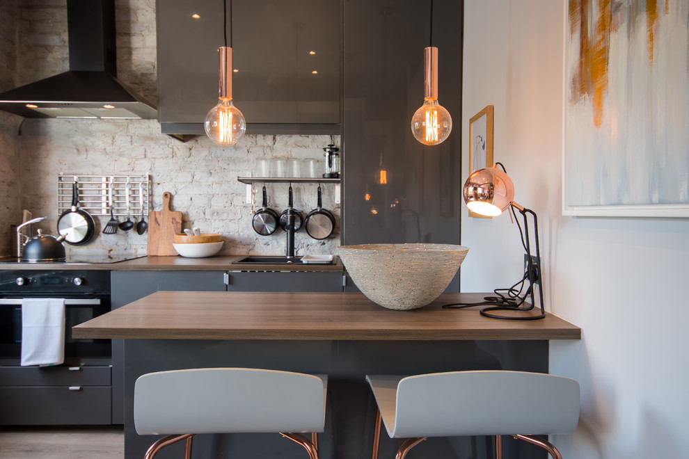 This is an example of a small contemporary kitchen in London with flat-panel cabinets, grey cabinets, brick splashback, a peninsula, brown benchtop, a drop-in sink, wood benchtops and black appliances.