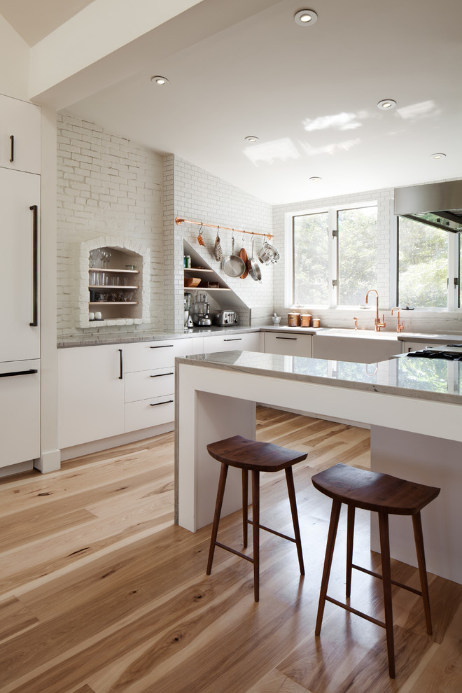 This is an example of a mid-sized contemporary kitchen in Boston with flat-panel cabinets, white cabinets, a farmhouse sink, panelled appliances, light hardwood floors and a peninsula.
