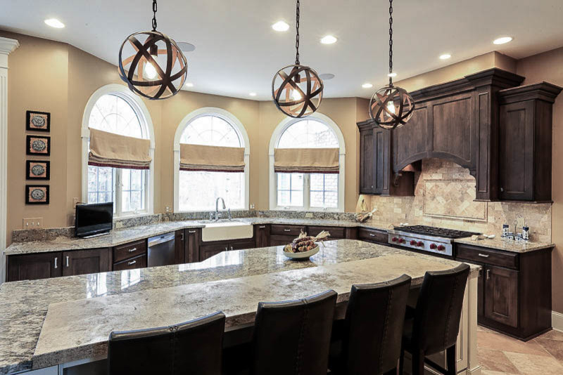 Example of a large mountain style u-shaped travertine floor eat-in kitchen design in Cleveland with a farmhouse sink, recessed-panel cabinets, dark wood cabinets, granite countertops, stone tile backsplash, stainless steel appliances, an island, beige backsplash and beige countertops