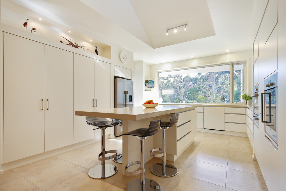 Photo of a contemporary u-shaped eat-in kitchen in Melbourne with flat-panel cabinets, white cabinets, window splashback, stainless steel appliances, with island, beige floor and beige benchtop.