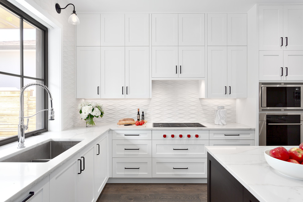 This is an example of a large transitional l-shaped kitchen in Vancouver with a double-bowl sink, recessed-panel cabinets, white cabinets, quartz benchtops, white splashback, porcelain splashback, stainless steel appliances, medium hardwood floors, with island, brown floor and white benchtop.