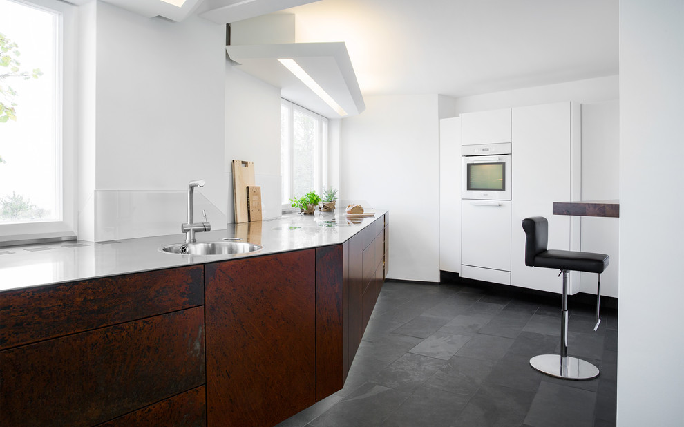 Photo of a mid-sized industrial l-shaped kitchen in Stuttgart with a drop-in sink, flat-panel cabinets, stainless steel appliances, slate floors and black floor.
