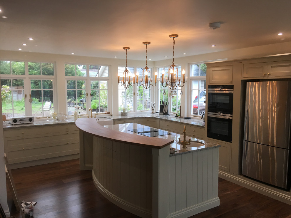 This is an example of a large contemporary eat-in kitchen in West Midlands with a farmhouse sink, shaker cabinets, beige cabinets, granite benchtops, stainless steel appliances, medium hardwood floors, with island, brown floor and beige benchtop.
