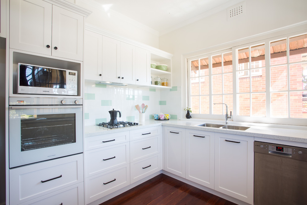 This is an example of a mid-sized traditional u-shaped separate kitchen in Brisbane with a double-bowl sink, shaker cabinets, white cabinets, quartz benchtops, green splashback, subway tile splashback, white appliances, dark hardwood floors, no island, red floor and white benchtop.