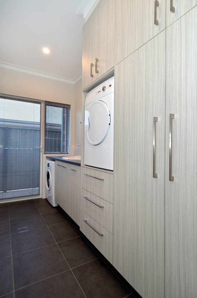 This is an example of a large contemporary single-wall dedicated laundry room in Other with a drop-in sink, flat-panel cabinets, light wood cabinets, laminate benchtops, grey walls, ceramic floors and a stacked washer and dryer.
