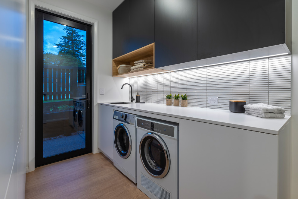 Design ideas for a contemporary single-wall laundry room in Auckland with an undermount sink, white cabinets, quartz benchtops, white splashback, mosaic tile splashback, laminate floors, a side-by-side washer and dryer, white benchtop, white walls and brown floor.