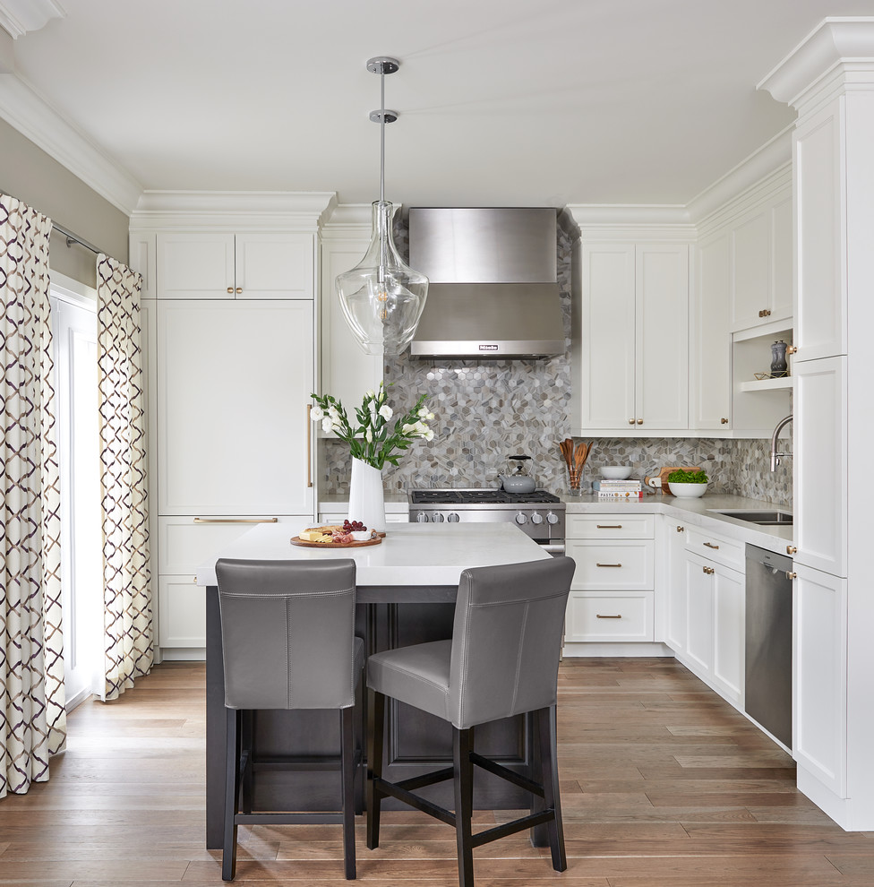This is an example of a transitional l-shaped kitchen in Toronto with an undermount sink, shaker cabinets, white cabinets, grey splashback, mosaic tile splashback, panelled appliances, medium hardwood floors, with island, brown floor and white benchtop.