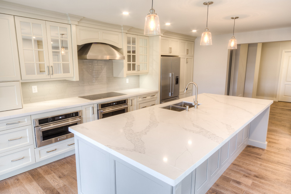 Open concept kitchen - mid-sized transitional galley light wood floor and beige floor open concept kitchen idea in Toronto with a double-bowl sink, recessed-panel cabinets, white cabinets, solid surface countertops, white backsplash, porcelain backsplash, stainless steel appliances, an island and white countertops