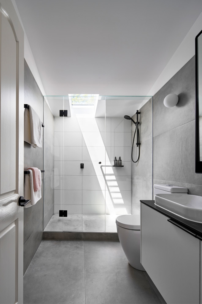 Contemporary bathroom in Melbourne with flat-panel cabinets, grey cabinets, a one-piece toilet, gray tile, ceramic tile, grey walls, ceramic floors, a vessel sink, solid surface benchtops, grey floor, a hinged shower door, black benchtops, a single vanity, a floating vanity and vaulted.