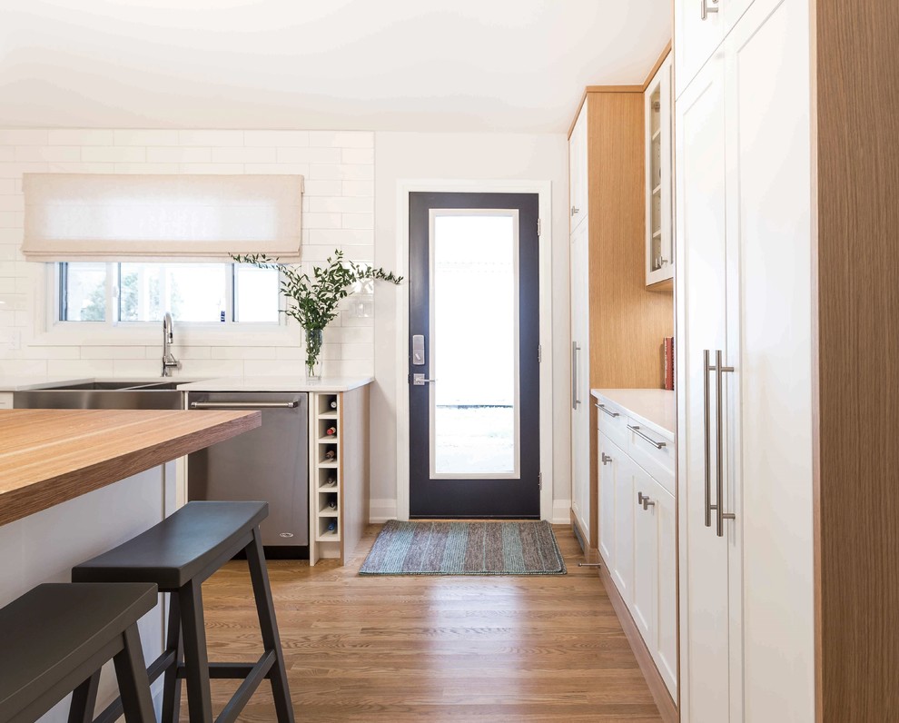 Small midcentury l-shaped open plan kitchen in Toronto with a farmhouse sink, recessed-panel cabinets, white cabinets, solid surface benchtops, white splashback, subway tile splashback, stainless steel appliances, light hardwood floors and with island.